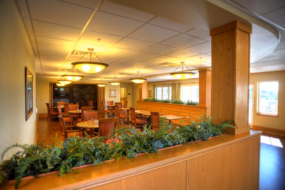 Well-lit communal dining room with wooden tables and chairs, pendant lights, and a planter divider.