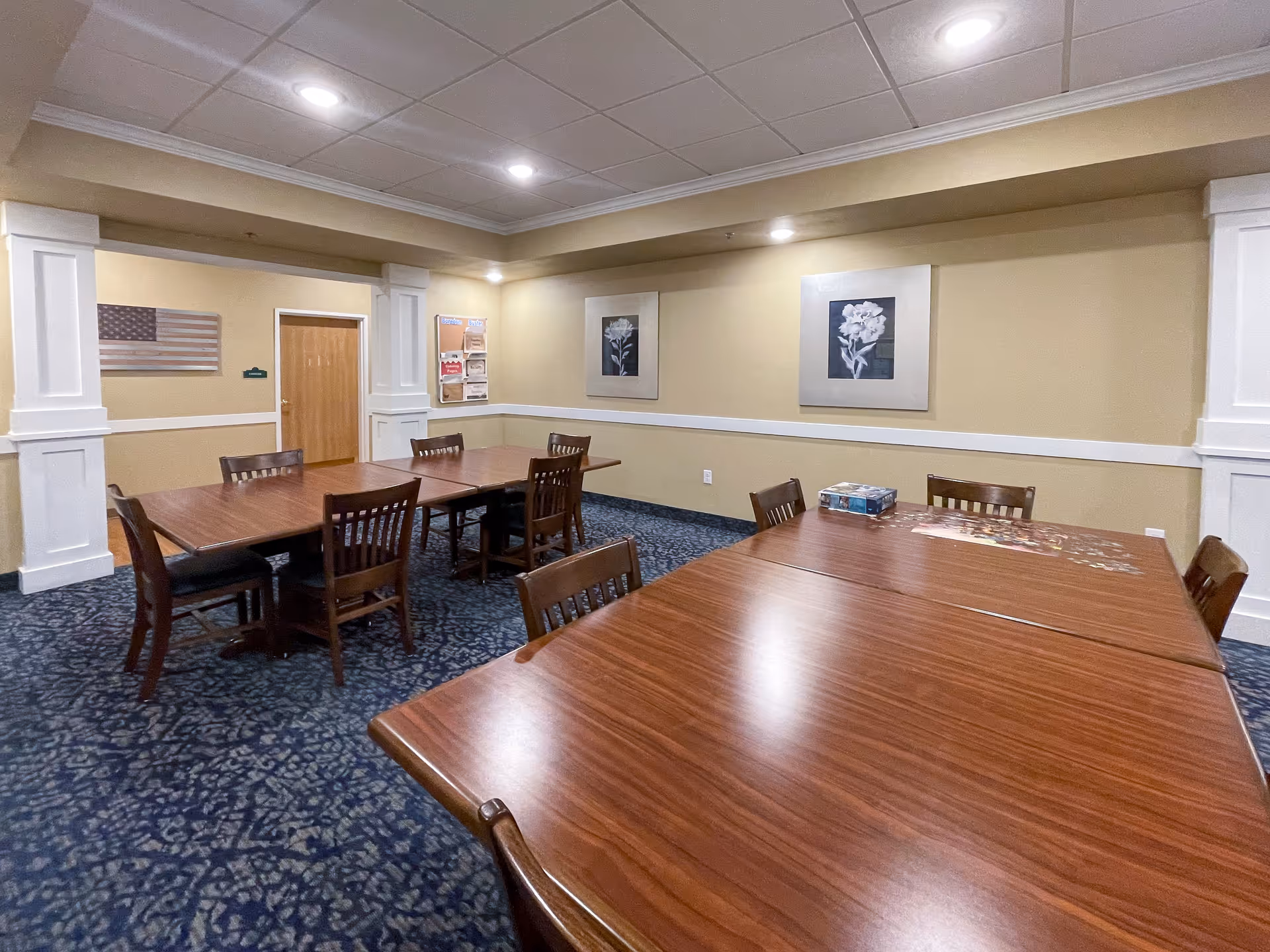 A well-lit room with beige walls and blue patterned carpet featuring two sets of wooden tables and chairs arranged for group seating. The walls are decorated with framed floral artwork and an American flag. There is a door and a bulletin board on one wall.