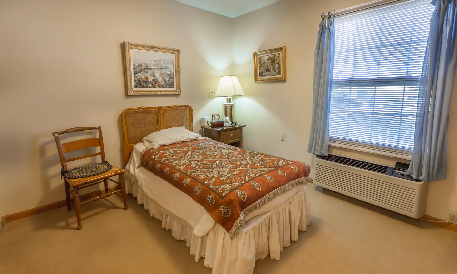 A small bedroom with a single bed covered with a patterned red and beige blanket. There is a wooden chair with a woven seat to the left of the bed. A nightstand with a lamp, tissue box, and small decorative items is next to the bed. Two framed paintings hang on the walls, and a window with blue curtains and blinds is on the right side of the room.