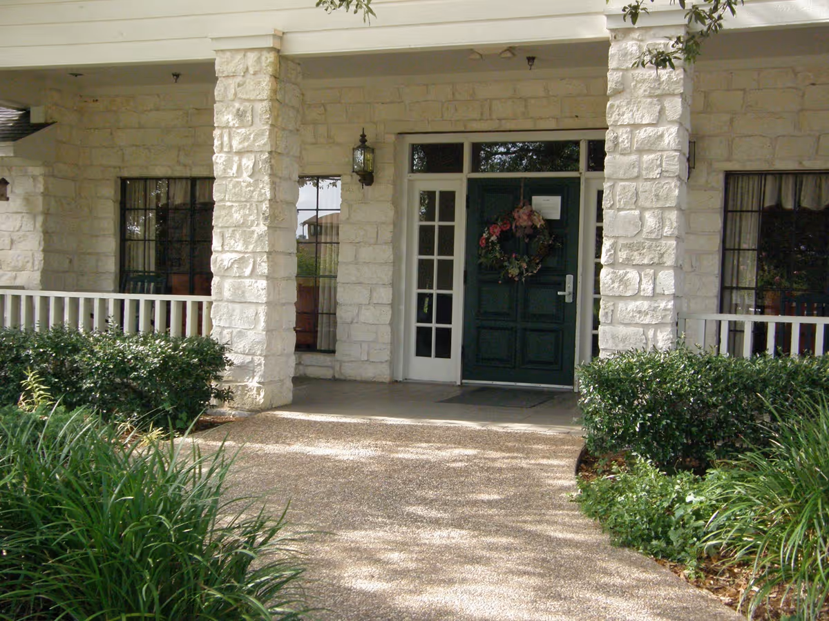 Entrance to a building with a green door decorated with a floral wreath, flanked by white stone pillars and windows with black frames. There are bushes and plants along the walkway leading to the door.