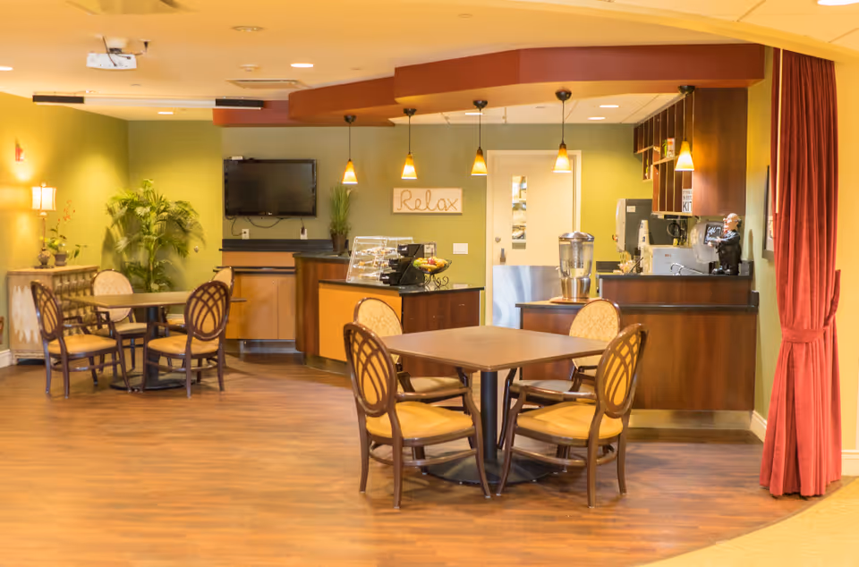 A cozy senior living common area with wooden floors, green walls, and warm lighting. There are two tables each surrounded by four cushioned chairs. A small counter area with pendant lights hangs above, featuring a water dispenser, a small display case, and a TV mounted on the wall. The word 'Relax' is displayed on a sign above the counter. A red curtain is partially visible on the right side of the image.