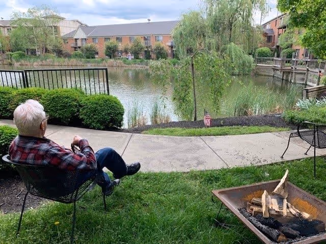 An elderly person with white hair and glasses sits on a metal chair facing a pond surrounded by greenery and residential buildings. There is a fire pit with wood in the foreground and a paved walkway along the pond's edge.