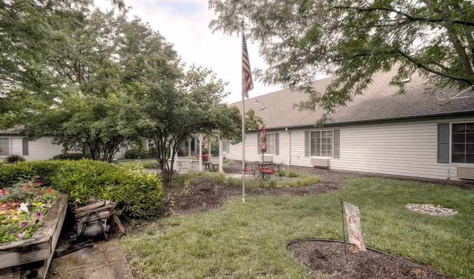 Outdoor garden area at Vintage Park at Lenexa featuring green grass, trees, bushes, a flower bed, a flagpole with an American flag, a small seating area with a table and chairs, and a light-colored building with windows in the background.