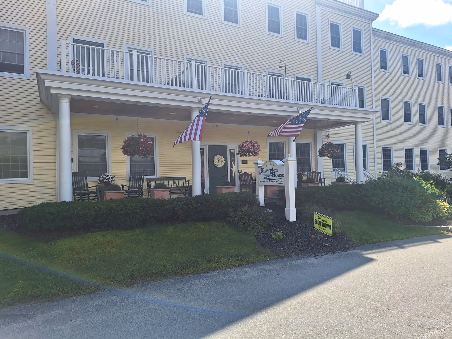 Exterior view of Riverglen House, a yellow multi-story building with white trim. The entrance features a covered porch with white columns, hanging flower baskets, rocking chairs, and two American flags. There is a sign in front of the porch that reads 'Riverglen House' and a smaller yellow sign on the lawn that says 'Please Slow Down'.