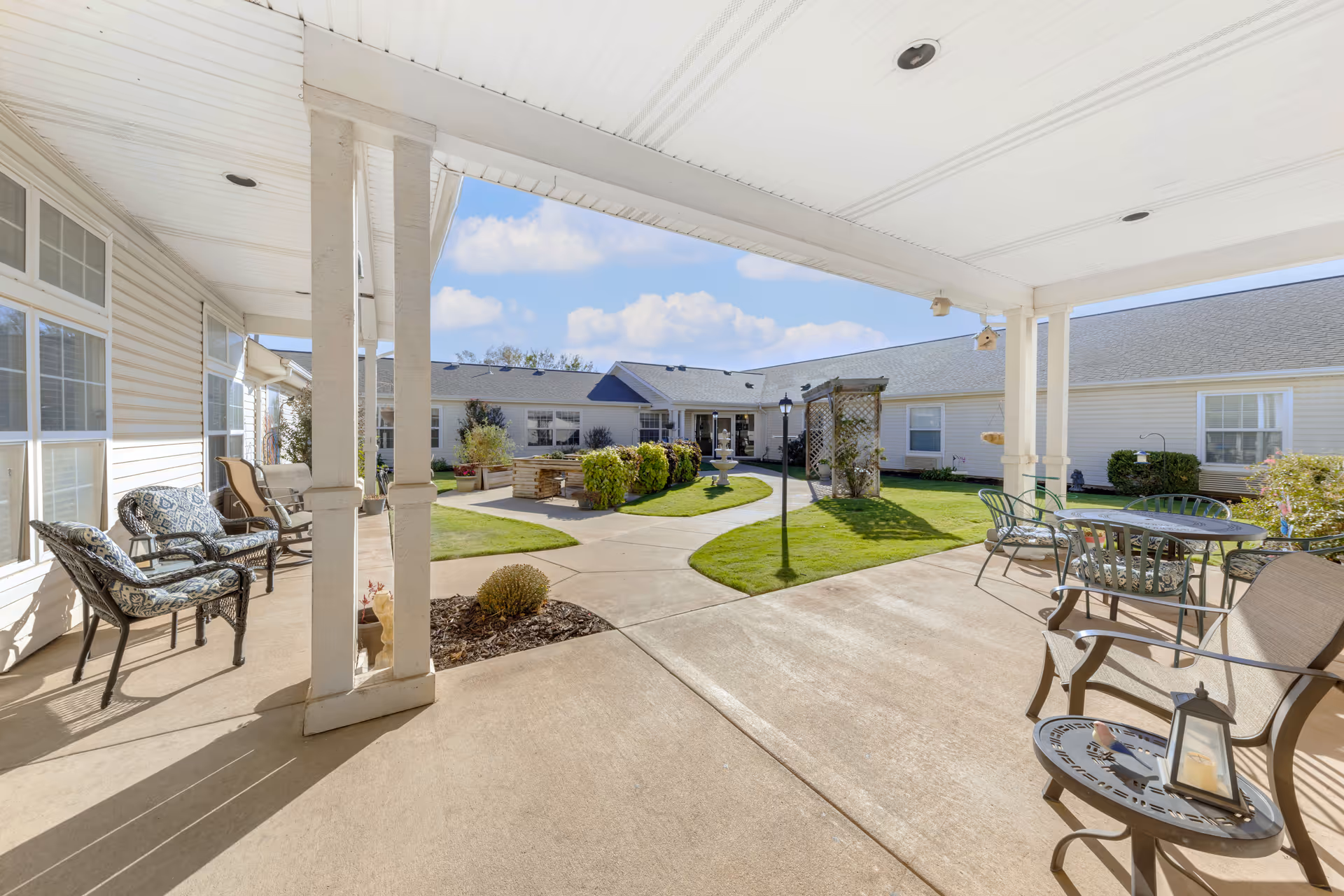 Covered patio area with outdoor seating including cushioned chairs and a table with a lantern, overlooking a courtyard with a walking path, green lawn, plants, a fountain, and a trellis. The courtyard is surrounded by a single-story building with white siding and multiple windows under a blue sky with some clouds.