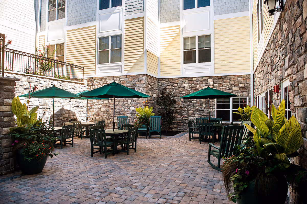 Outdoor courtyard area with stone walls and paved floor, featuring several green tables and chairs with green umbrellas. Large potted plants with green and yellow leaves are placed around the seating area. The courtyard is surrounded by a multi-story building with beige and gray siding and multiple windows.