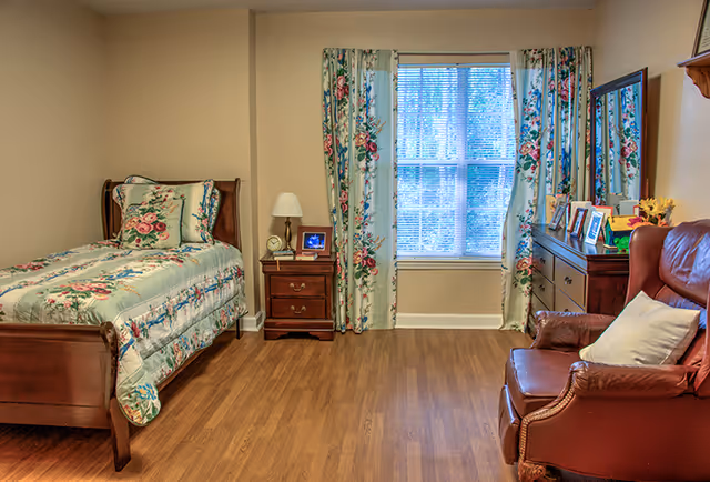 A cozy bedroom with a wooden single bed covered in floral bedding, a matching nightstand with a lamp and clock, floral curtains framing a window, a wooden dresser with framed photos and decorations, and a brown leather armchair with a white pillow.