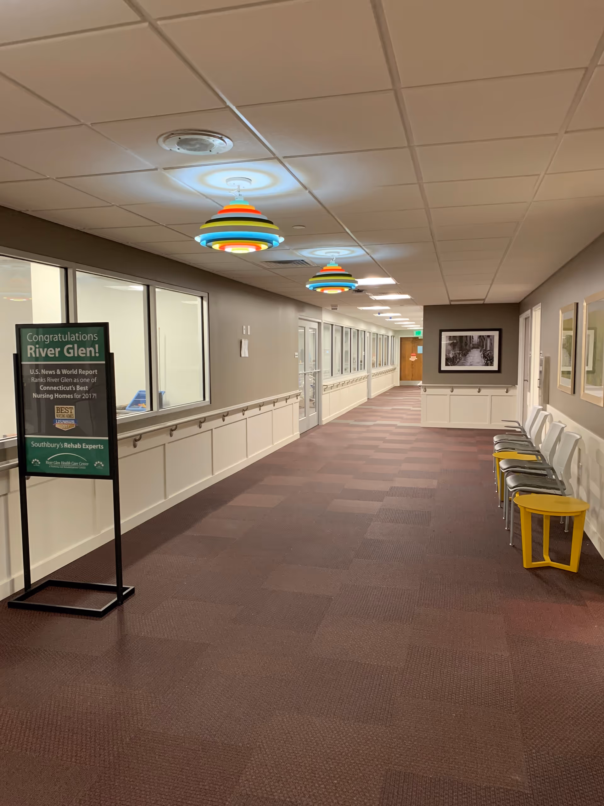 A long, well-lit hallway in a healthcare facility with brown carpeted floor and beige walls. On the left side, there are large windows and a sign congratulating River Glen for being ranked one of Connecticut's best nursing homes. On the right side, there are white chairs with a yellow side table and framed pictures on the wall. Colorful pendant lights hang from the ceiling.