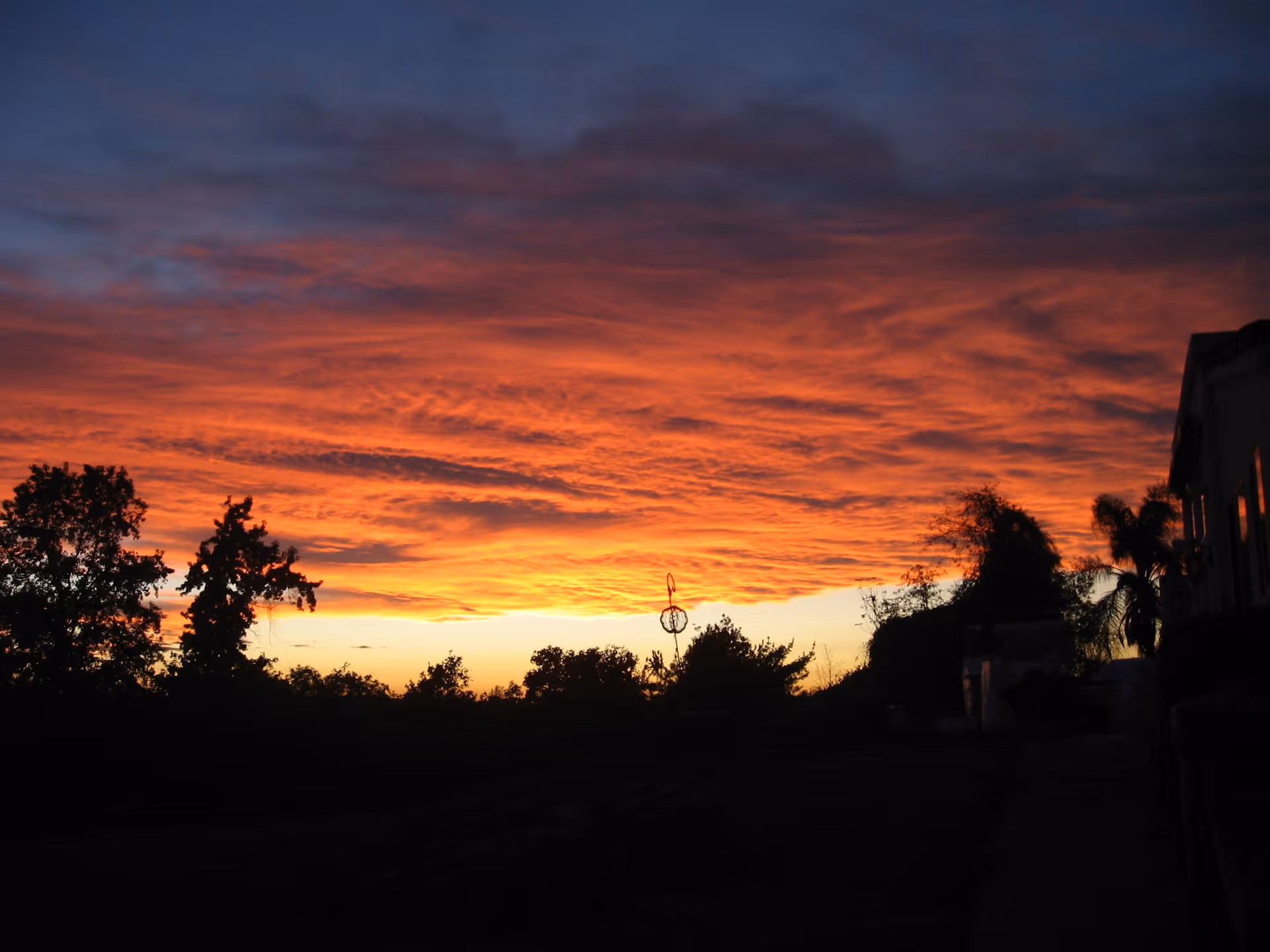 A vibrant sunset with a sky filled with orange and red clouds above a silhouette of trees and a building on the right side.