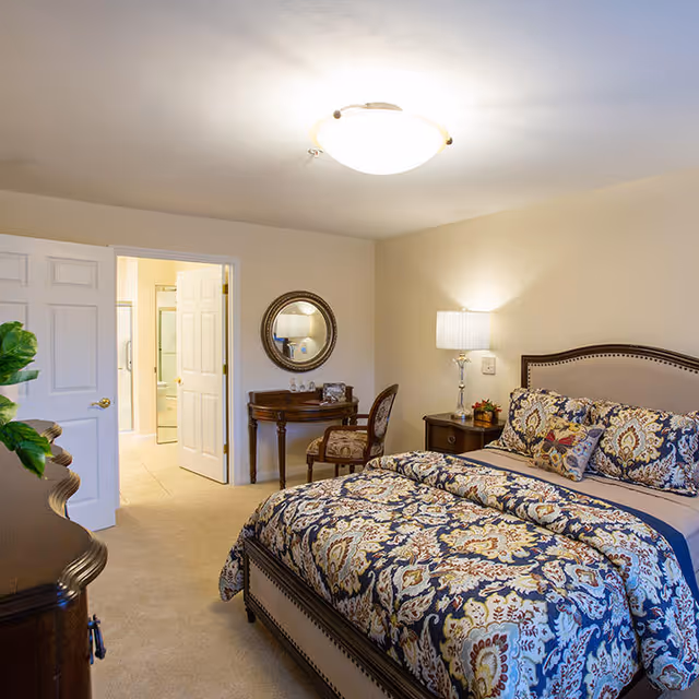 Well-lit furnished bedroom with a patterned bed, nightstand, lamp and an open door leading to an en-suite bathroom.