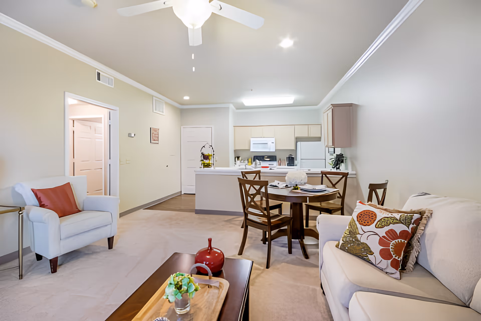 Interior view of a senior living facility apartment at Elison Independent Living of Statesman Club featuring a living room with a white armchair and sofa with decorative pillows, a wooden coffee table with a plant and decorative items, a round dining table with four chairs, and a kitchen area with white cabinets, a refrigerator, microwave, and stove in the background.