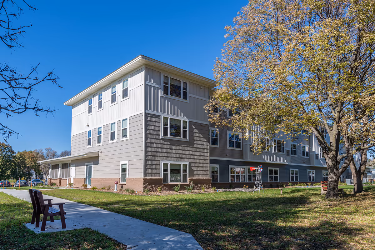 Exterior view of a three-story senior living building with a sidewalk, benches, and trees on a sunny day.