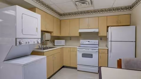 Compact kitchen with stacked washer/dryer, stove, sink, fridge, and wooden cabinets.