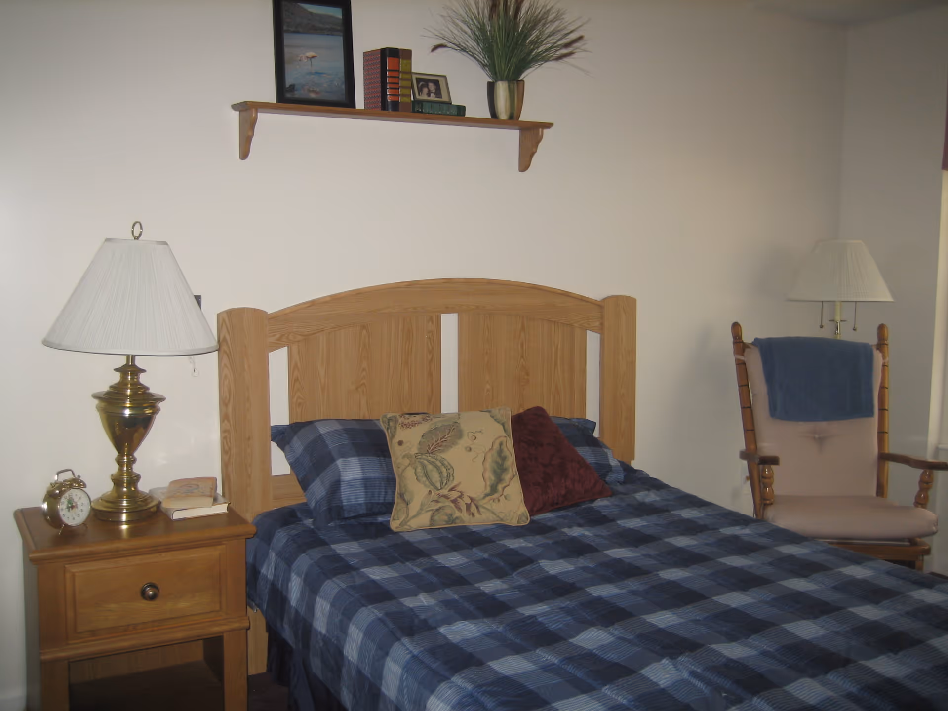 Bedroom with a wooden headboard, blue plaid bedspread, nightstand with a lamp and alarm clock, and a rocking chair.