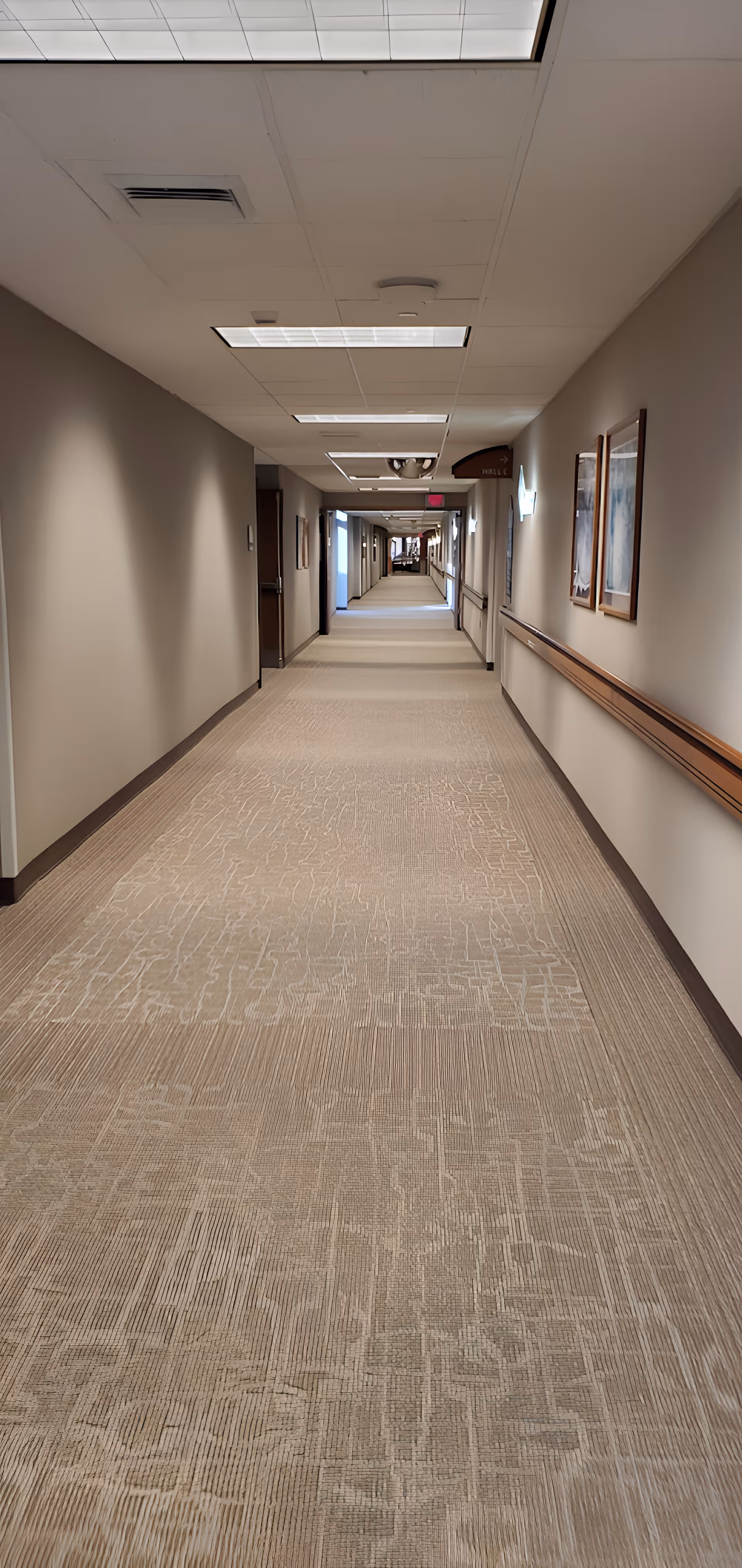 A long, well-lit hallway in a medical or rehabilitation facility with beige walls and carpeted floor. The ceiling has recessed lighting panels, and there are handrails along the walls. Several framed pictures are hung on the right wall, and doors line the hallway on both sides. A sign indicating 'Hall C' is visible on the right side.