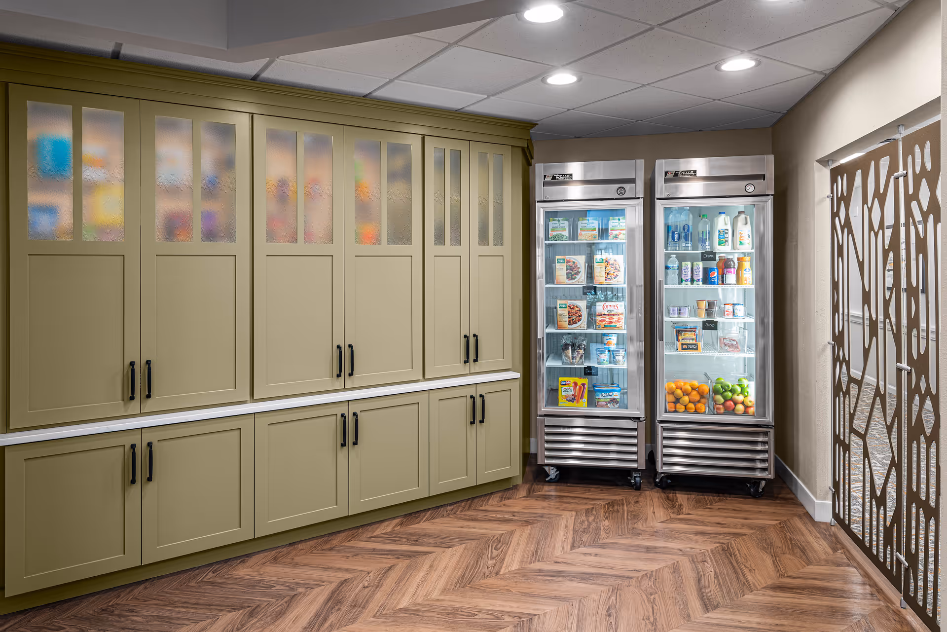Interior view of a pantry area with olive green cabinets on the left and two glass-door refrigerators stocked with beverages, fruits, and food items on the right. The floor has a wood herringbone pattern and there is a decorative metal partition on the far right.