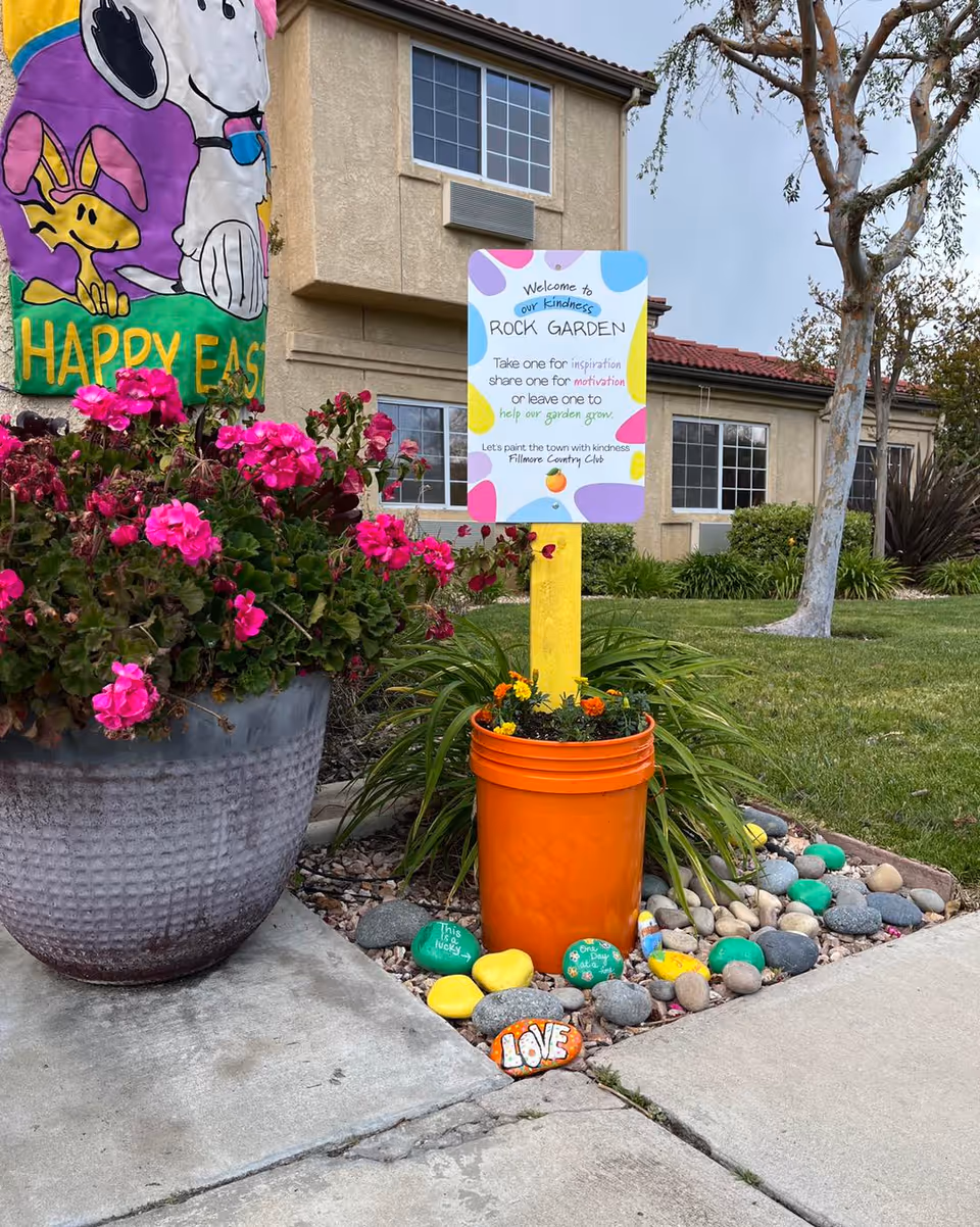 Outdoor garden area at Fillmore Country Club featuring a large planter with bright pink flowers, a colorful sign reading 'Welcome to our kindness ROCK GARDEN' with instructions to take, share, or leave rocks for inspiration and motivation, and an orange bucket with small flowers surrounded by painted rocks with positive messages. A building with beige walls and windows is visible in the background along with a tree and green lawn.