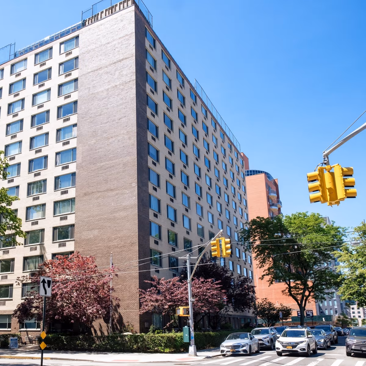Exterior view of a multi-story residential building on a sunny day with clear blue sky. The building has many windows and is surrounded by trees with green and reddish leaves. Several cars are parked along the street, and traffic lights and street signs are visible at the intersection.