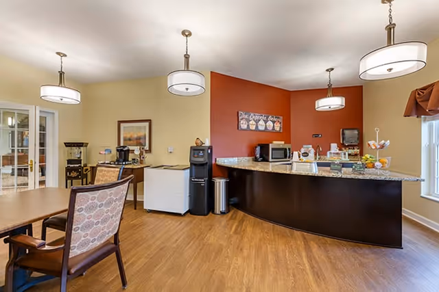 Communal dining and refreshment area with a curved granite service counter, coffee station, chairs, and pendant lighting.