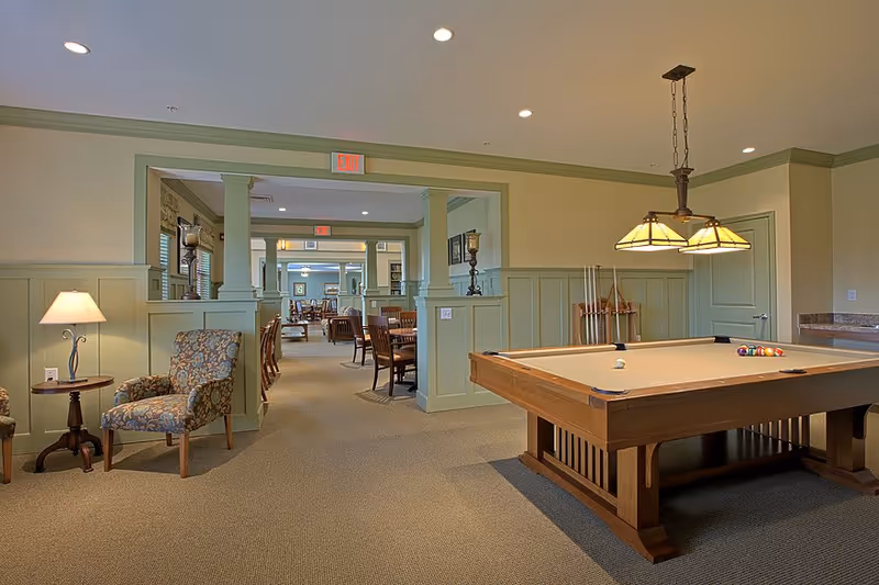 Interior view of a senior living facility recreation room featuring a pool table with balls and cues, a hanging light fixture above it, and a seating area with floral upholstered chairs and a side table with a lamp. Beyond the room, there is a dining area with wooden tables and chairs, soft green wainscoting on the walls, and recessed ceiling lights.