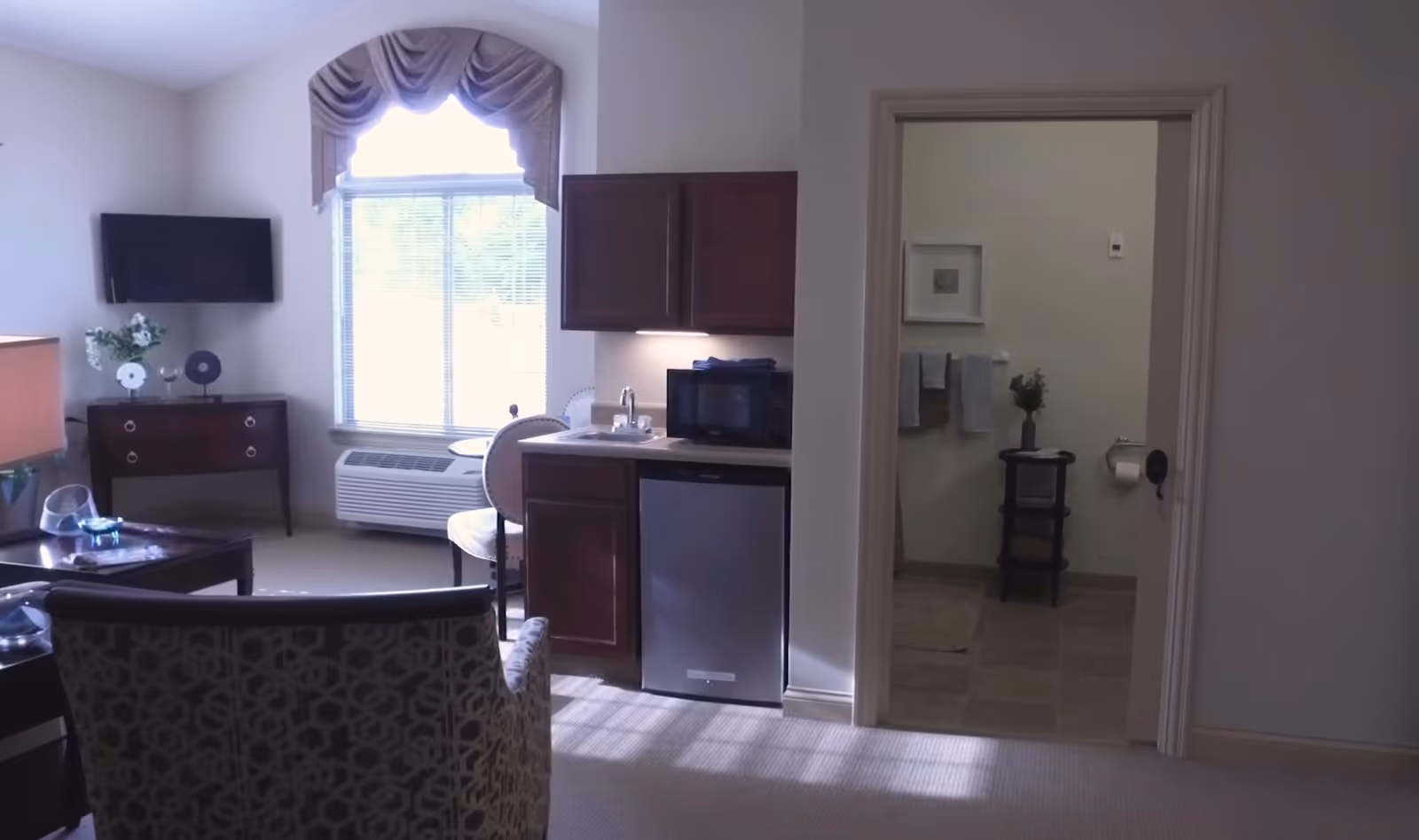 Small senior living studio interior with seating area, kitchenette and an open bathroom doorway.