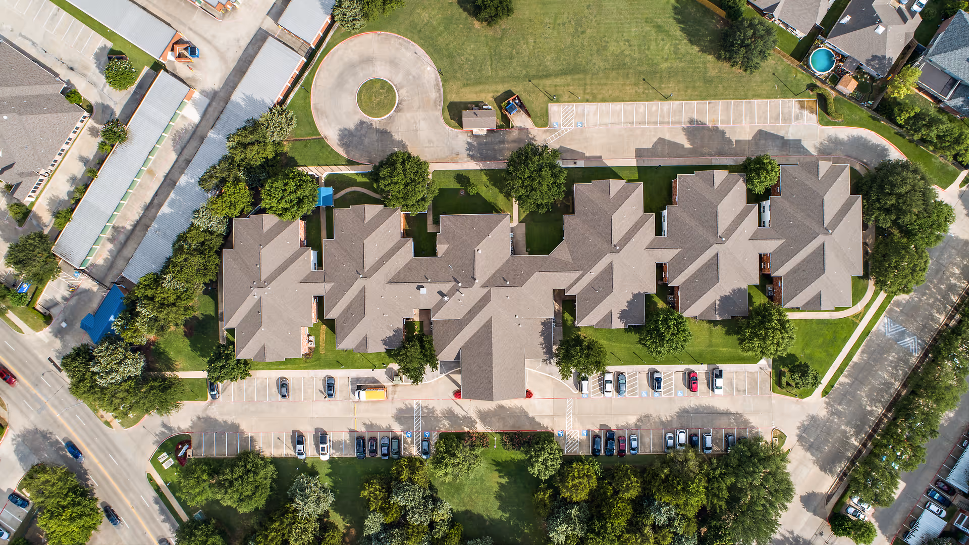 Aerial view of Morada Grand Prairie facility showing multiple connected buildings with brown roofs, surrounded by green lawns, trees, and parking lots with several cars parked. The facility is bordered by roads and neighboring properties.