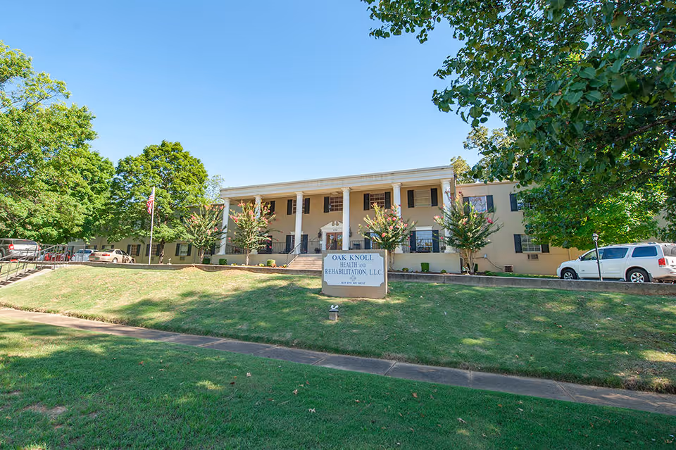Front exterior view of Oak Knoll Health and Rehabilitation, LLC building with a lawn and trees in the foreground, several cars parked on the sides, and a clear blue sky.