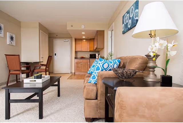 Bright living room with a brown sofa and blue pillows, coffee table, side table with lamp and a small dining area and kitchenette in the background.