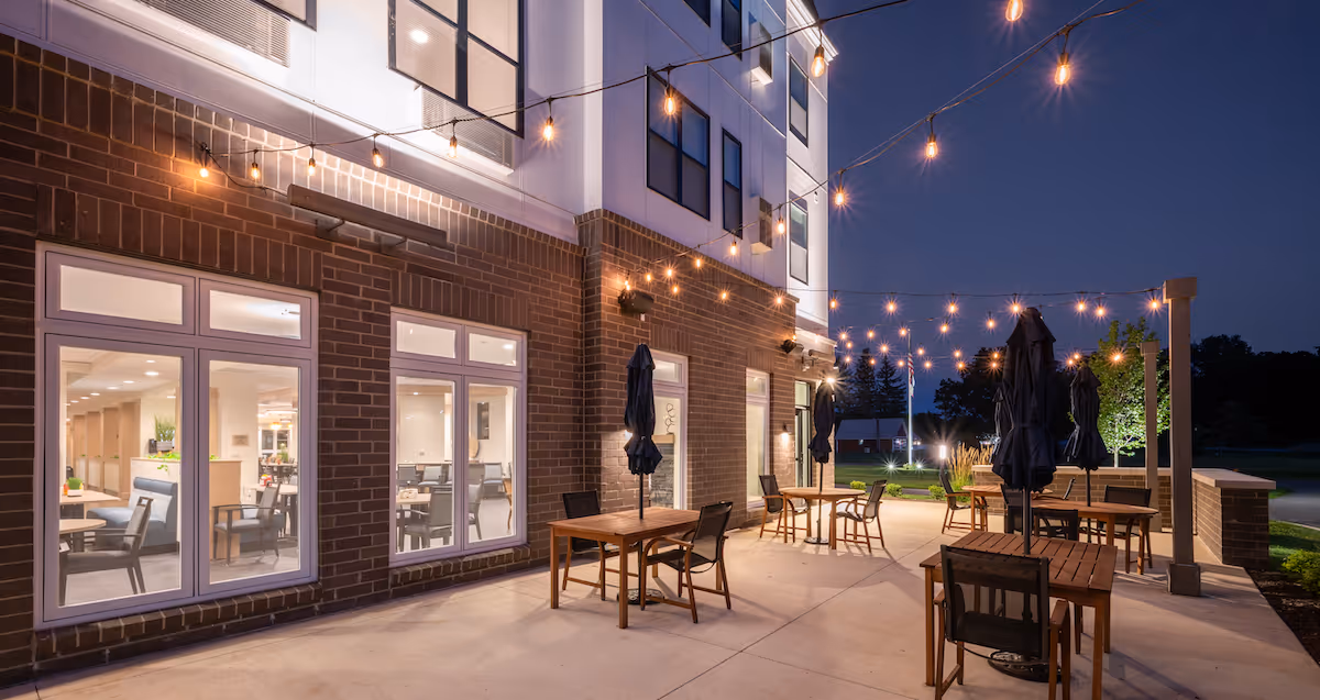 Outdoor patio area at night with string lights hanging above wooden tables and chairs, some tables have closed umbrellas. The building has large windows showing a well-lit interior dining area.