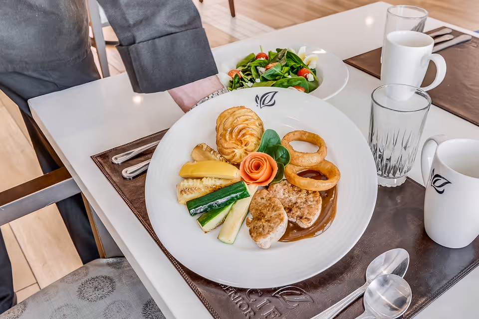A plated meal on a white table in a dining setting, featuring grilled vegetables, two pieces of meat with sauce, two onion rings, a pastry, and a garnish shaped like a rose. There is also a bowl of salad, a glass of water, and a white mug on the table, with brown placemats and silverware.