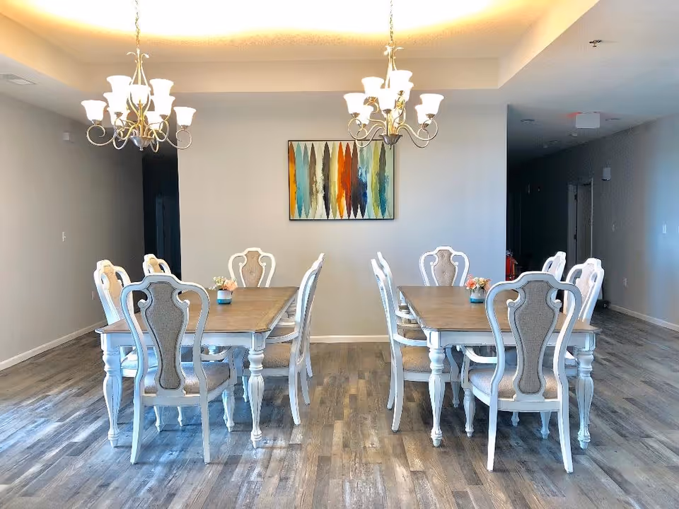 A bright dining room with two wooden dining tables, each surrounded by six white chairs with beige cushioned backs and seats. Two elegant chandeliers hang from the ceiling above the tables. A colorful abstract painting is centered on the wall behind the tables. The floor is covered with wood-patterned flooring, and the walls are painted light gray.