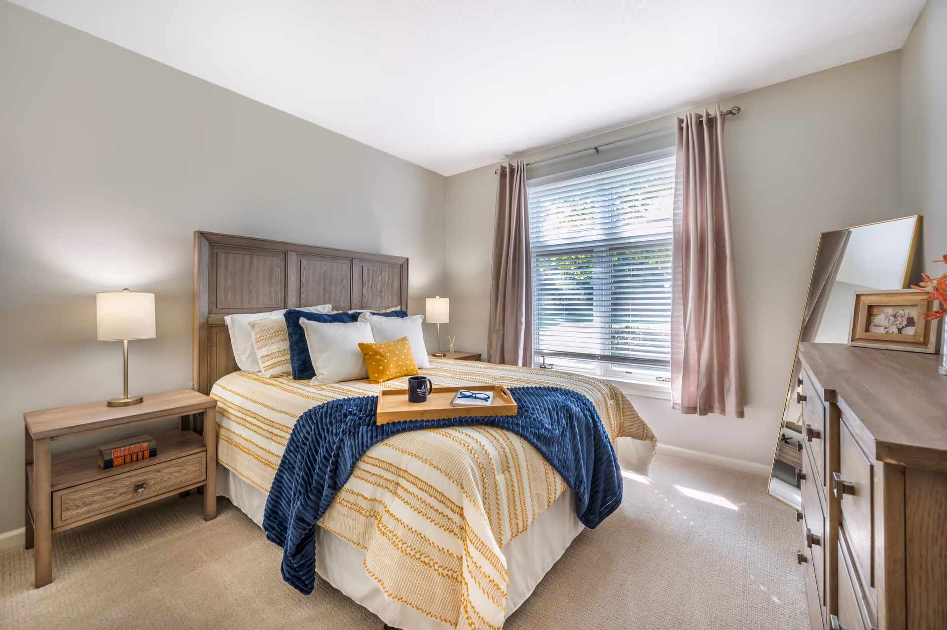 A bright and cozy bedroom with a wooden bed frame and matching nightstand and dresser. The bed is made with a white and yellow striped comforter, multiple pillows, and a blue throw blanket. A wooden tray with a mug and glasses rests on the bed. There are two lamps on either side of the bed, a large window with blinds and pink curtains, and a tall mirror leaning against the wall next to the dresser.