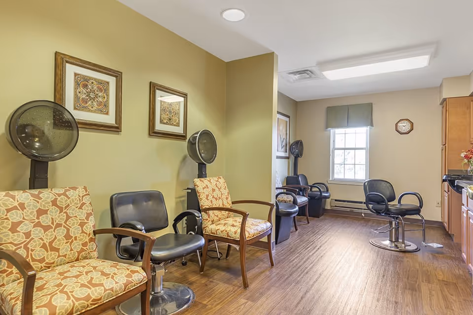 Interior view of a salon area in a senior living facility with several salon chairs and hair dryers along the walls. The room has wooden flooring, beige walls with framed artwork, a window with a green valance, and a clock on the wall.