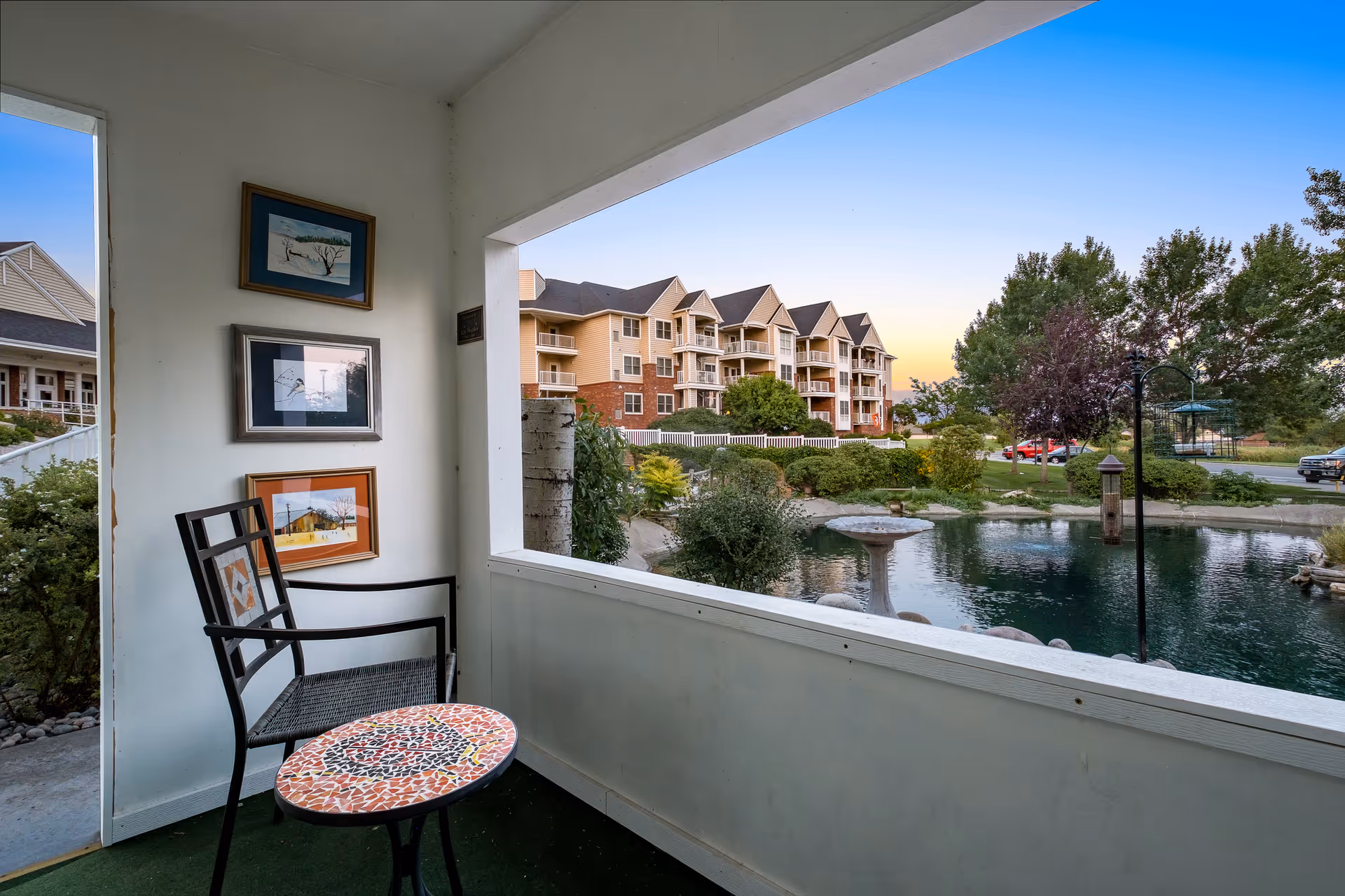 A covered porch area with a black metal chair and a small mosaic-topped table. Three framed pictures hang on the white wall. The porch overlooks a landscaped pond with a fountain, surrounded by trees and shrubs. In the background, there is a multi-story residential building with balconies under a clear sky at sunset.