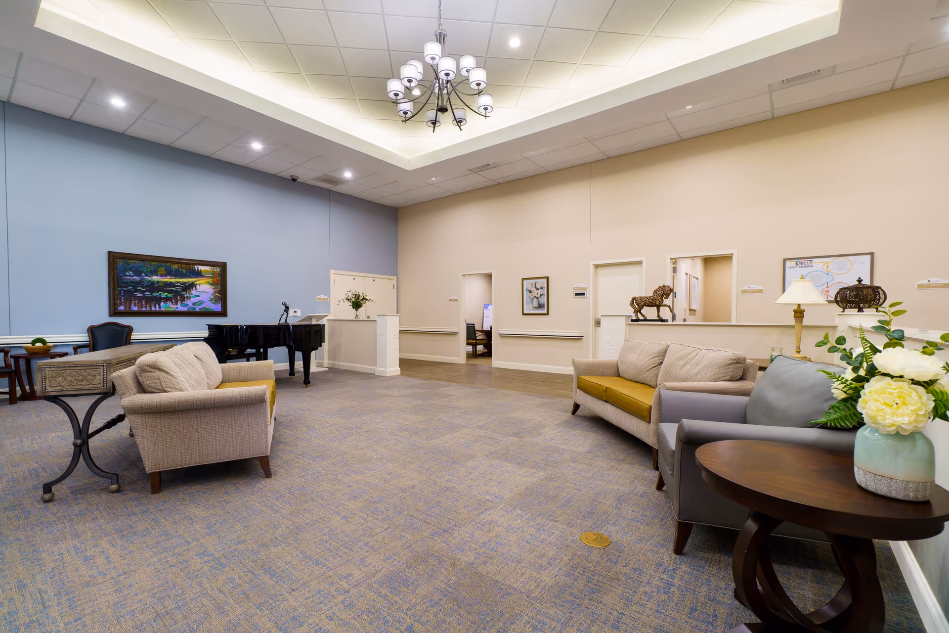 A spacious and well-lit common area in a senior living facility featuring comfortable seating including sofas and armchairs, a grand piano against a light blue wall, a chandelier hanging from the ceiling, framed artwork on the walls, and a side table with a vase of flowers.
