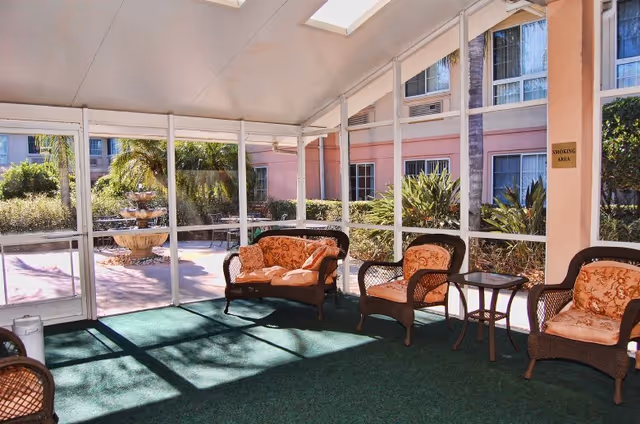 A screened-in porch area with wicker furniture including a loveseat and three chairs with floral cushions, a small table, and a green carpet. Outside the porch, there is a multi-tiered water fountain and lush greenery with palm trees and bushes. The building exterior is visible with pink walls and multiple windows.