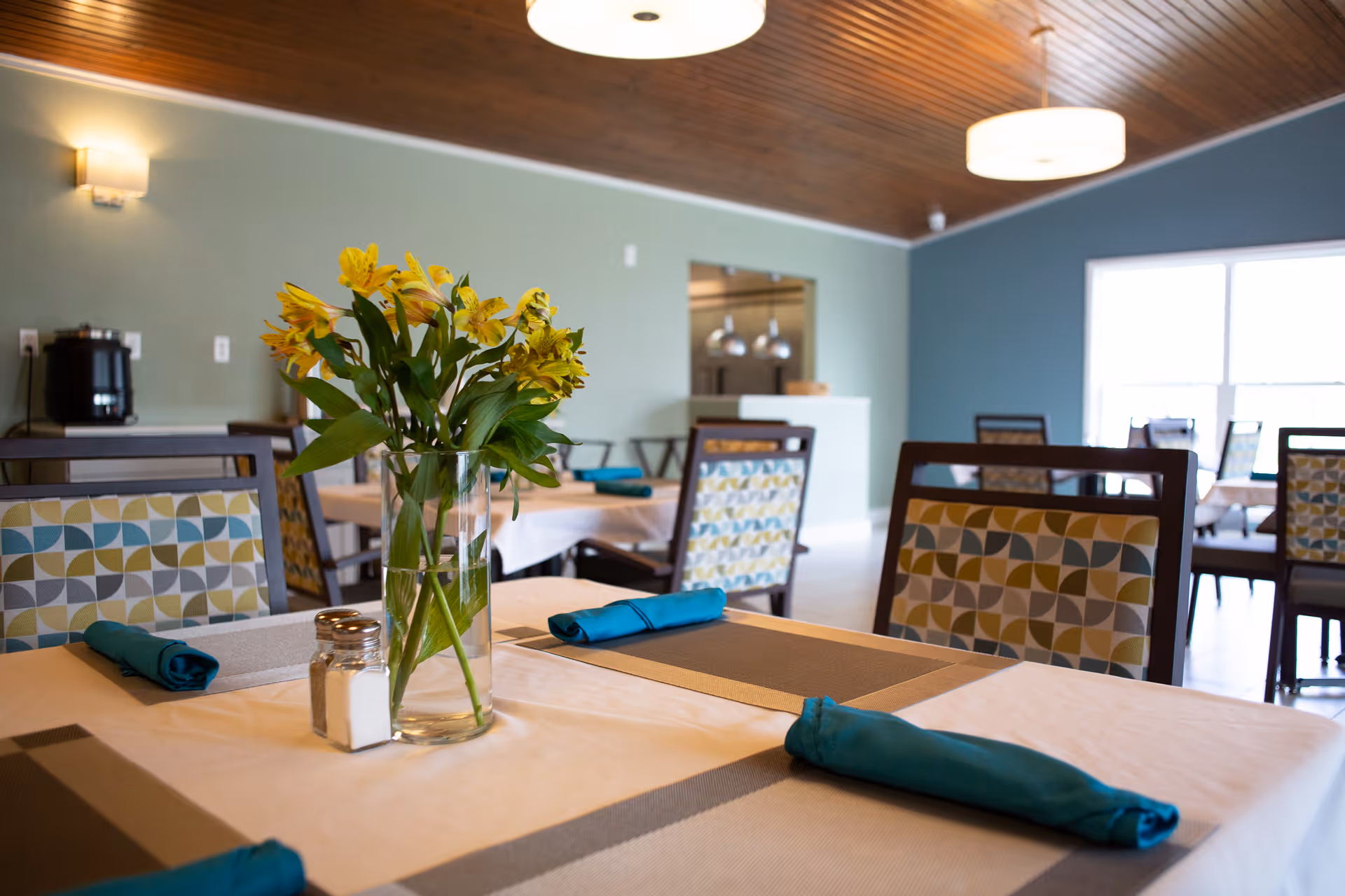 Dining room with set tables featuring a vase of yellow flowers, teal napkins, patterned chairs, and pendant lights.