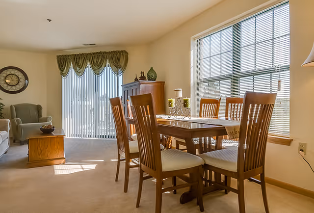 Sunlit dining area with a wooden table and chairs next to a living room with armchairs and sliding glass doors.