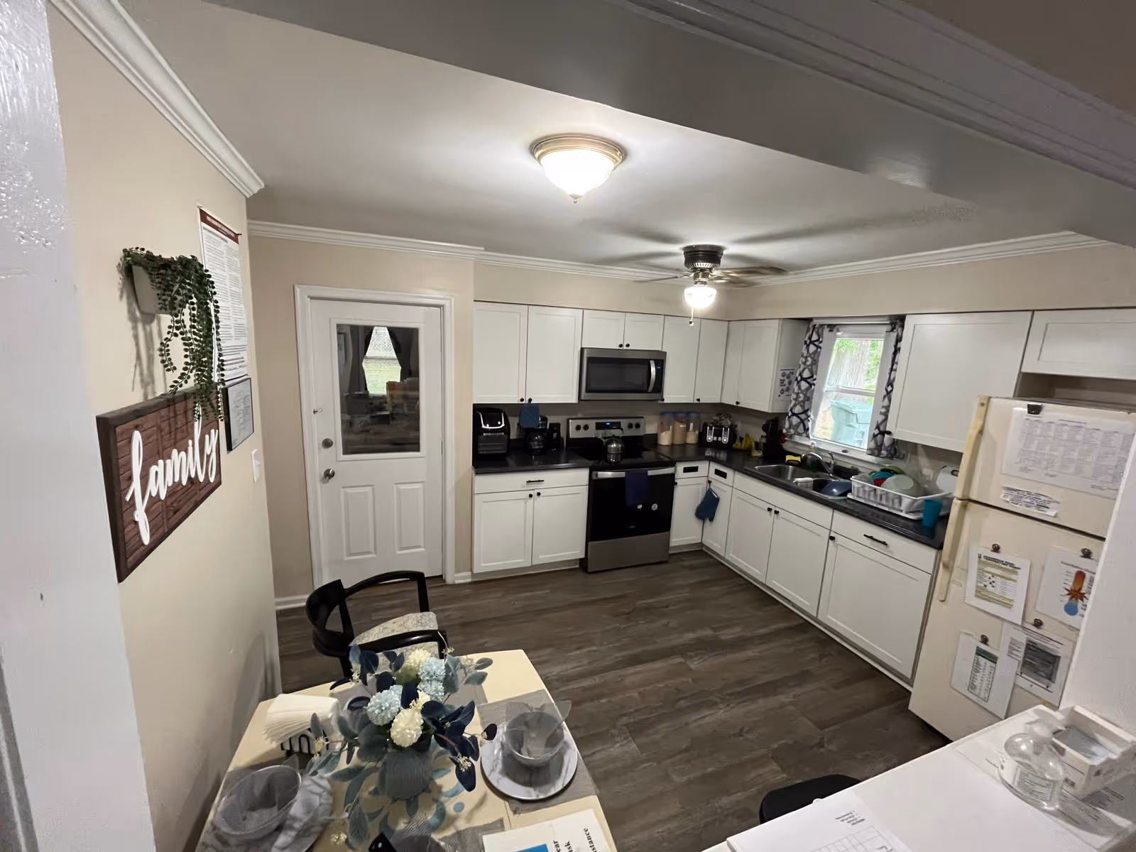 A clean and modern kitchen with white cabinets, stainless steel stove and microwave, a white refrigerator with papers attached, a double sink with dishes in a drying rack, and a small dining table set with plates and a floral centerpiece. There is a door with a window, a window with patterned curtains, and a wall decoration that says 'family'.