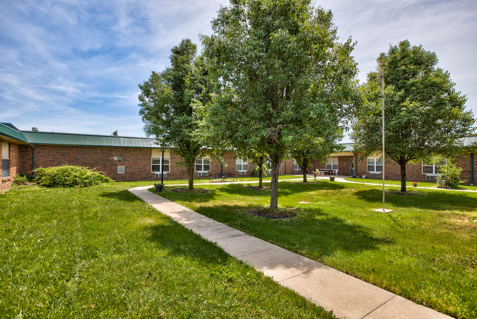 A grassy courtyard area with a concrete walkway leading through it, surrounded by several leafy green trees and a brick building with multiple windows in the background under a partly cloudy sky.