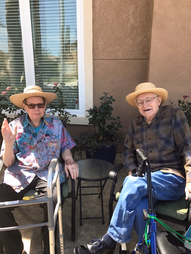 Two elderly men sitting outside on patio chairs next to a small glass table. Both are wearing straw hats and glasses. One man is wearing a colorful patterned shirt and has a walker in front of him, while the other man is wearing a plaid shirt and jeans with a blue walker beside him. There are plants and a window with blinds behind them.