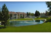 Two-story apartment buildings surrounding a landscaped pond with fountains and green lawns.