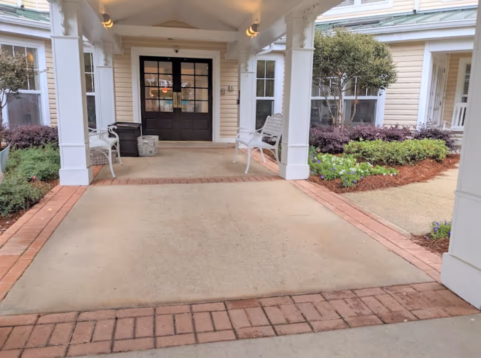 Covered entrance with a brick-trimmed concrete drive leading to double doors, benches, and landscaped planting beds.
