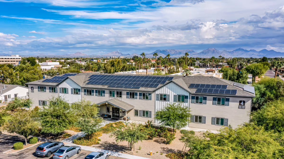 Aerial view of a two-story senior living facility building with solar panels on the roof, surrounded by trees and landscaping, with a parking lot in front and mountains visible in the background under a partly cloudy sky.
