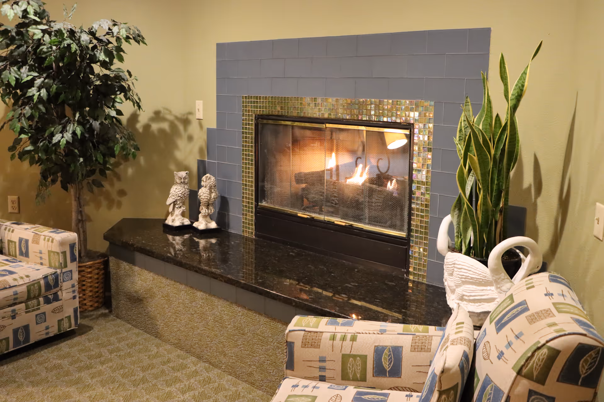 Cozy living room area with a lit fireplace surrounded by blue and mosaic tiles. Two patterned armchairs with leaf and geometric designs face the fireplace. There are decorative owl statues on the black granite hearth, a potted leafy plant on the left, and a tall snake plant in a white swan planter on the right.