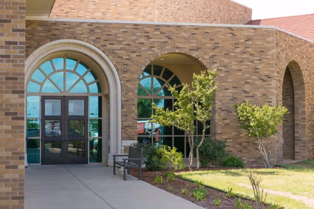 Brick building entrance with arched windows and double glass doors, a bench, and small landscaped trees.
