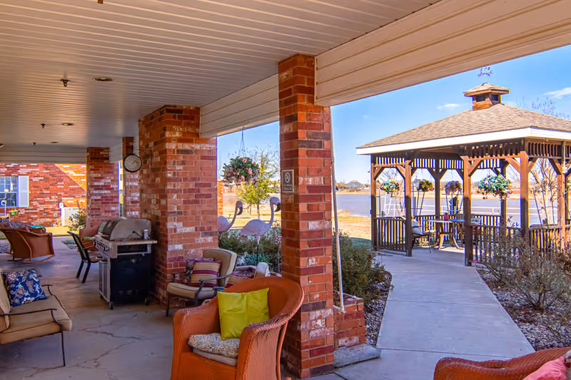 Covered outdoor patio area with brick pillars, wicker chairs with colorful cushions, a grill, and decorative flamingos. A concrete pathway leads to a wooden gazebo overlooking a lake with hanging flower baskets.