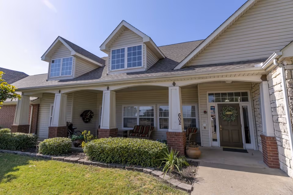 Front exterior view of a senior living facility building with beige siding, brick accents, a covered porch with seating, and a green door decorated with a wreath. The building number 602 is visible on a column. There are shrubs and a small garden in front of the porch under a clear blue sky.