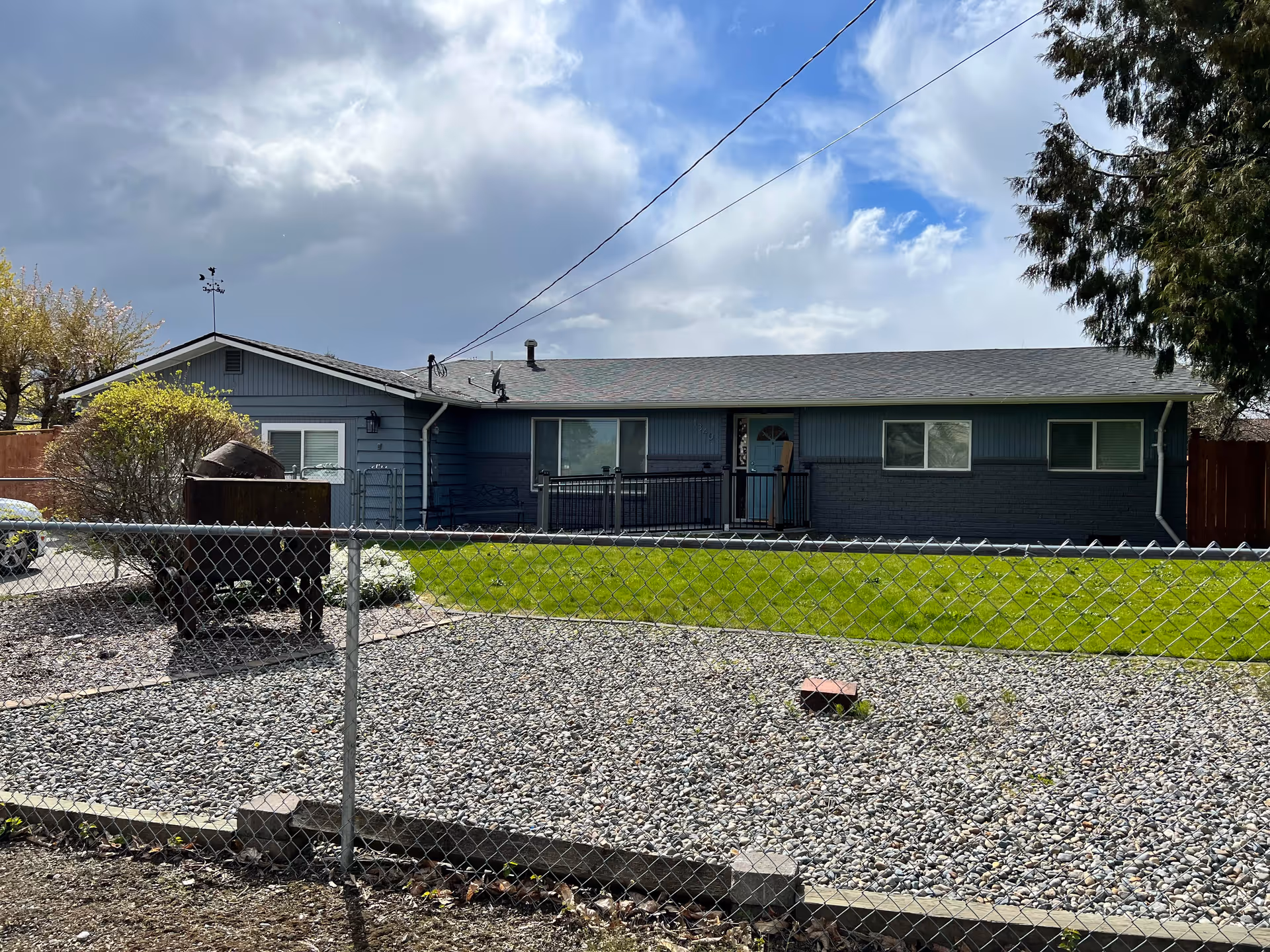 Single-story blue house with a fenced front yard featuring a green lawn and gravel area under a partly cloudy sky.