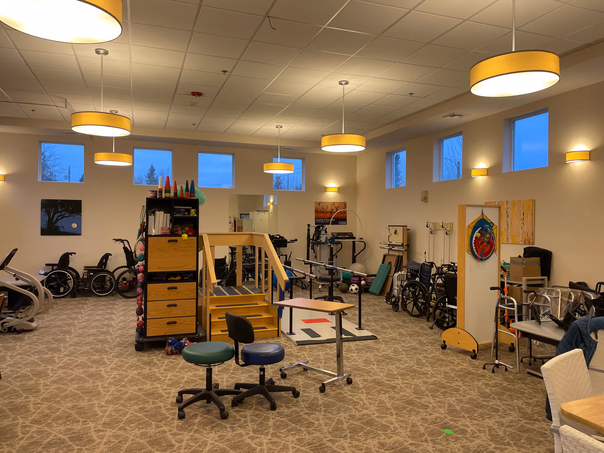 A spacious rehabilitation therapy room with exercise equipment, wheelchairs, therapy stairs, and stools beneath hanging pendant lights.